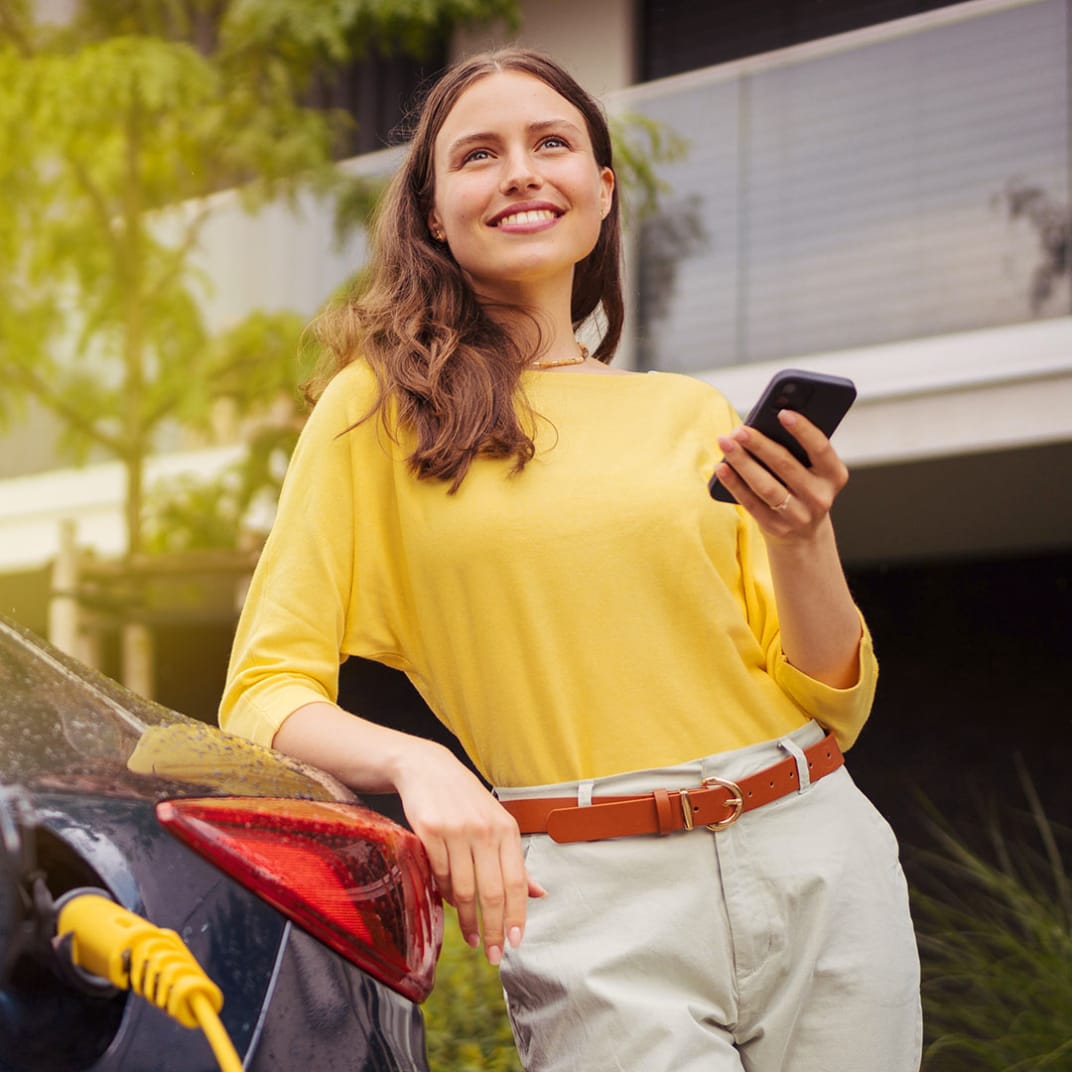 Frau steht mit Smartphone an einem E-Auto mit Ladekabel
