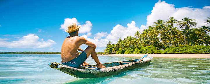 Person auf einem kleinen Boot im türkisfarbenen Meer vor einer tropischen Insel - symbolisches Motiv für Fernreisen und Urlaubsplanung