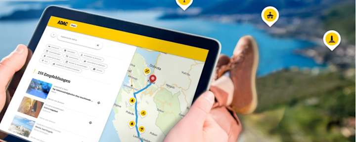 Tablet mit ADAC Routenplaner zeigt eine Reiseroute und Empfehlungen vor einer Küstenlandschaft