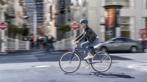 Eine Frau in Jeans, Lederjacke und mit Helm, fährt auf einem Herrenfahrrad durch die Stadt
