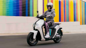 Ein weißer Elektro-Roller namens Neo von Yamaha fährt durch die Stadt an einer bunten Mauer entlang