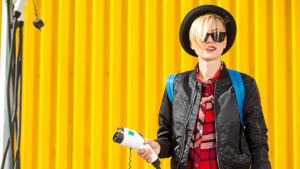 Junge Frau steht vor gelbem Container mit dem Ladekabel für ein Elektroauto in der Hand