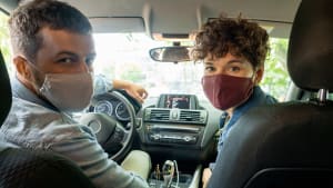 Mann und Frau mit Mundschutzmaske im Auto blicken nach hinten zur Rücksitzbank zum Betrachter