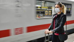 Frau steht mit Mundschutz zur Corona-Zeit am Bahnsteig vor Zug der Deutschen Bahn
