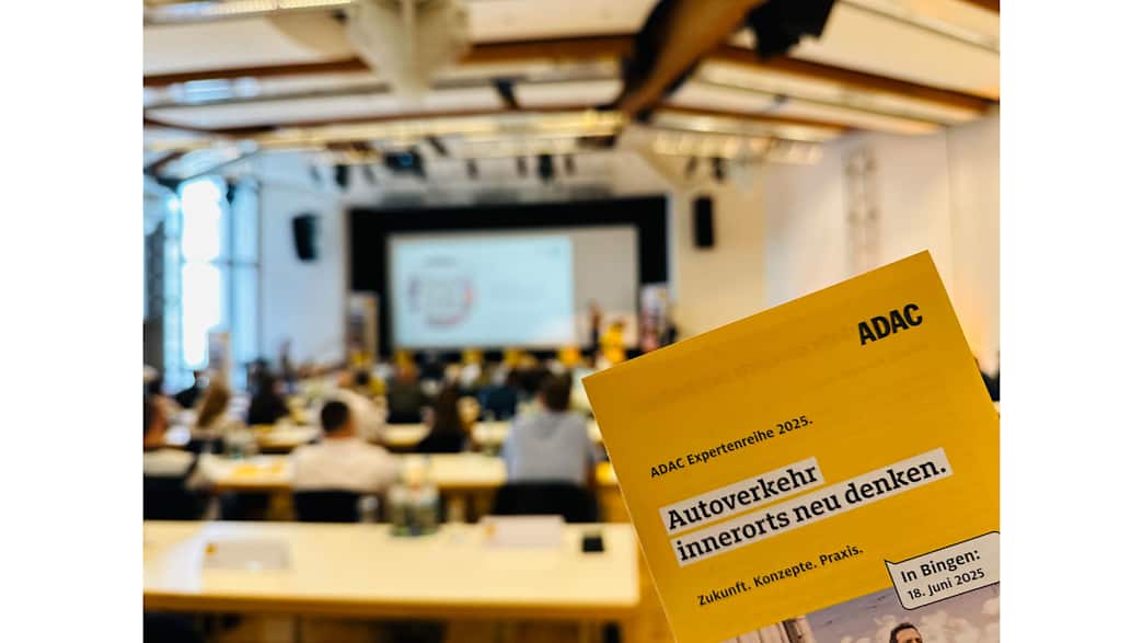 Eine Veranstaltung in einem großen Saal mit Publikum, im Vordergrund ist ein Flyer mit dem Titel „Autoverkehr innerorts neu denken“ zu sehen.