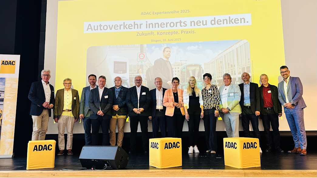 Eine Gruppe von Menschen steht vor einer Bühne unter einem Banner mit der Aufschrift „Autoverkehr innerorts neu denken“.