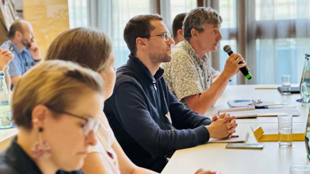 Teilnehmer einer Konferenz sitzen an einem Tisch und hören einer Person mit Mikrofon zu.