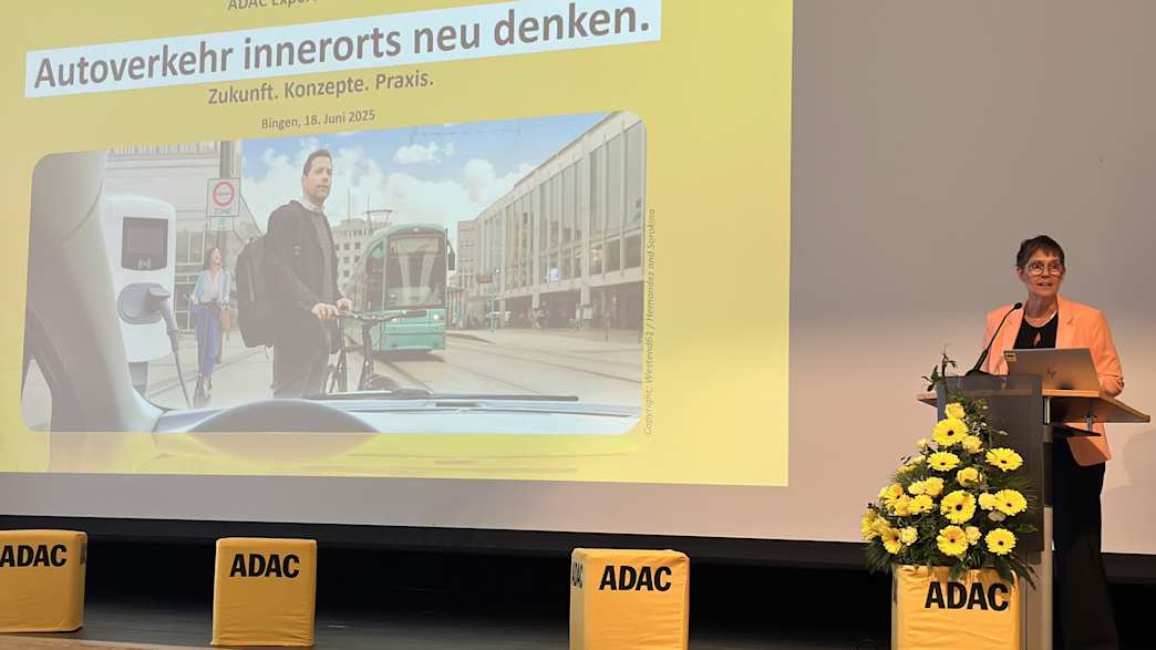 Eine Person am Rednerpult hält eine Präsentation auf der Bühne. Im Hintergrund ein großer Bildschirm mit dem Titel der Veranstaltung.