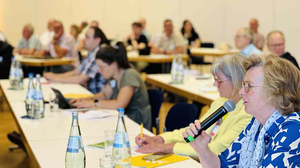 Eine Teilnehmerin stellt mit einem Mikrofon eine Frage, während sie an einem Tisch sitzt, umgeben von anderen Personen.