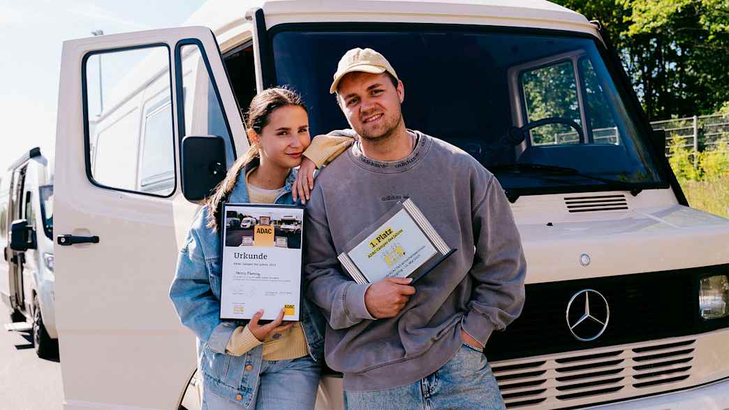 Ein Mann und eine Frau stehen lächelnd vor einem weißen Mercedes Wohnmobil. Sie halten eine Urkunde und eine Auszeichnung mit der Aufschrift "1. Platz ADAC Camper des Jahres".