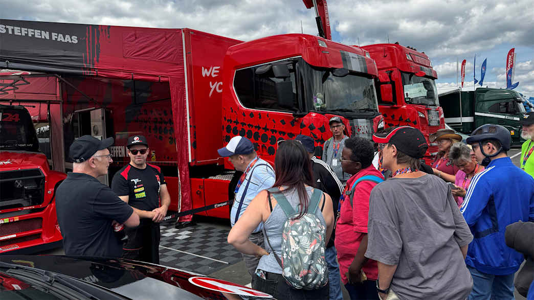 Eine Gruppe von Menschen steht vor roten Lastwagen auf einem Motorsport-Event. Einer der Menschen trägt ein schwarzes Hemd und spricht zu der Gruppe.