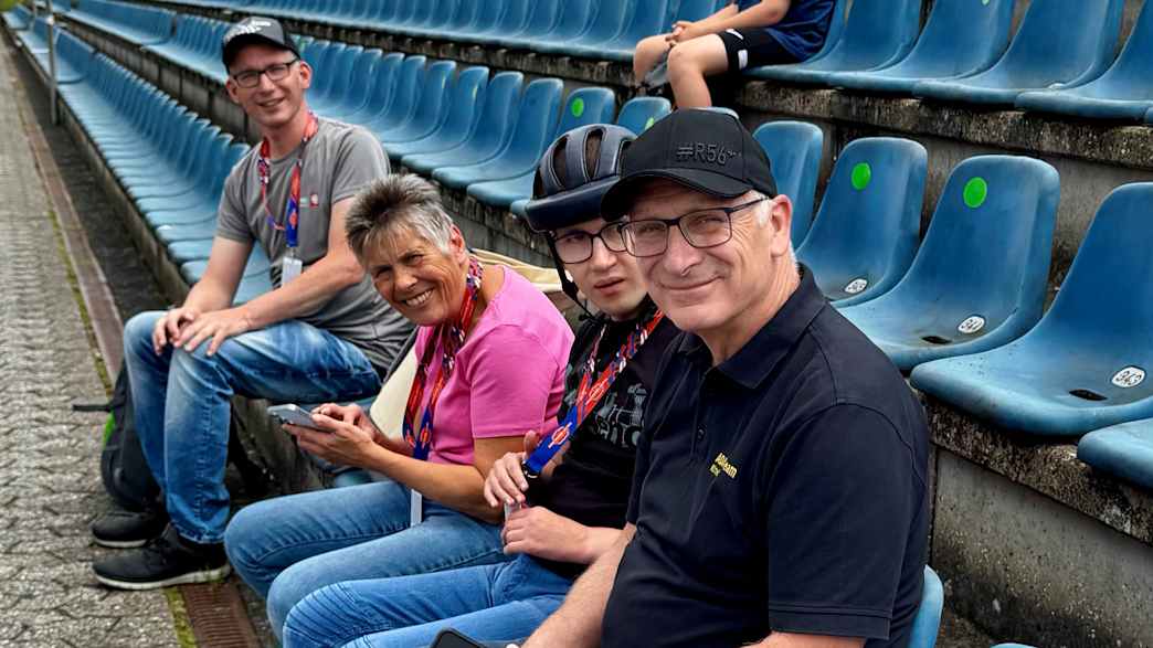 Vier Menschen sitzen auf blauen Tribünenplätzen. Sie lächeln und scheinen eine Veranstaltung zu beobachten.