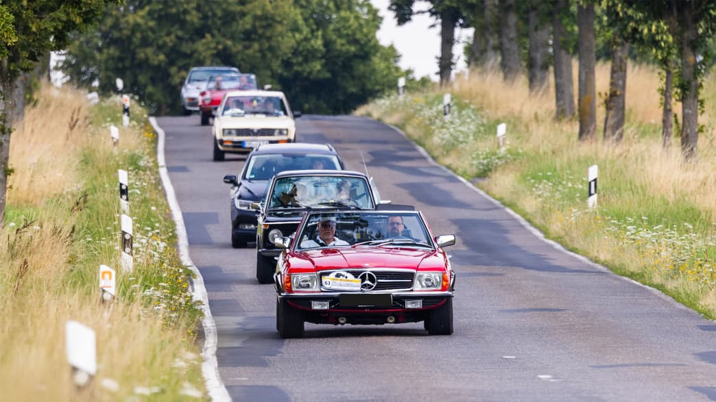Eine beeindruckende Kolonne klassischer Fahrzeuge mit einem roten Mercedes-Benz SL an der Spitze, der die Straße entlangfährt und für Fahrvergnügen und Qualität steht.