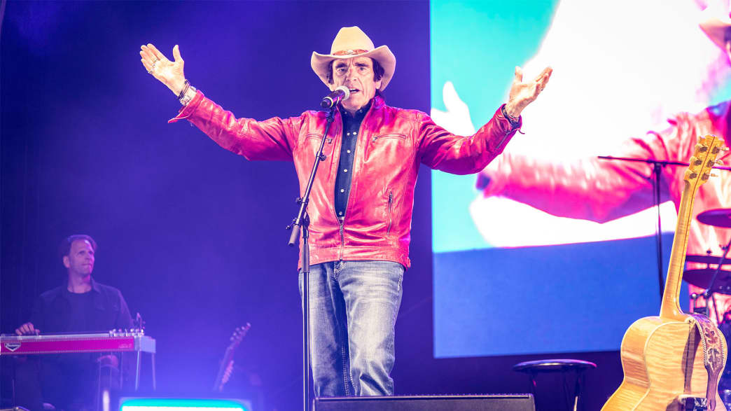 Country-Sänger, der live auf der Bühne auftritt, mit roter Lederjacke und Cowboyhut, vor einem lebhaften Publikum im Hintergrund.