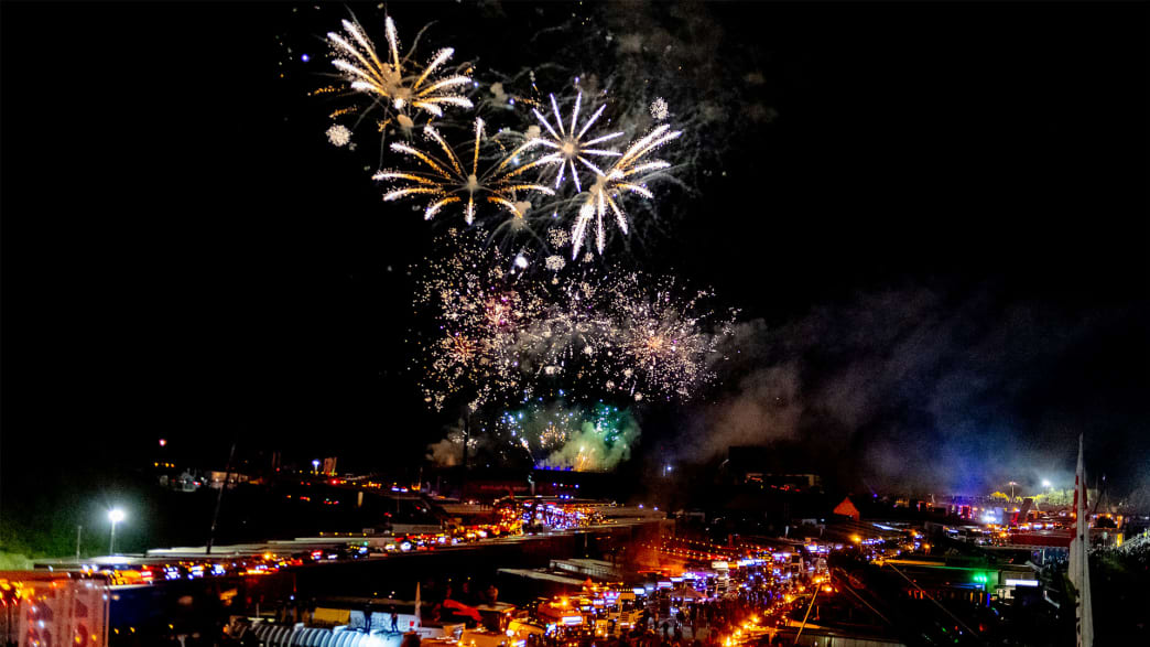 Spektakuläres Feuerwerk, das den Nachthimmel über einer geschäftigen Veranstaltung erleuchtet, mit beleuchteten Trucks und Teilnehmern unten.