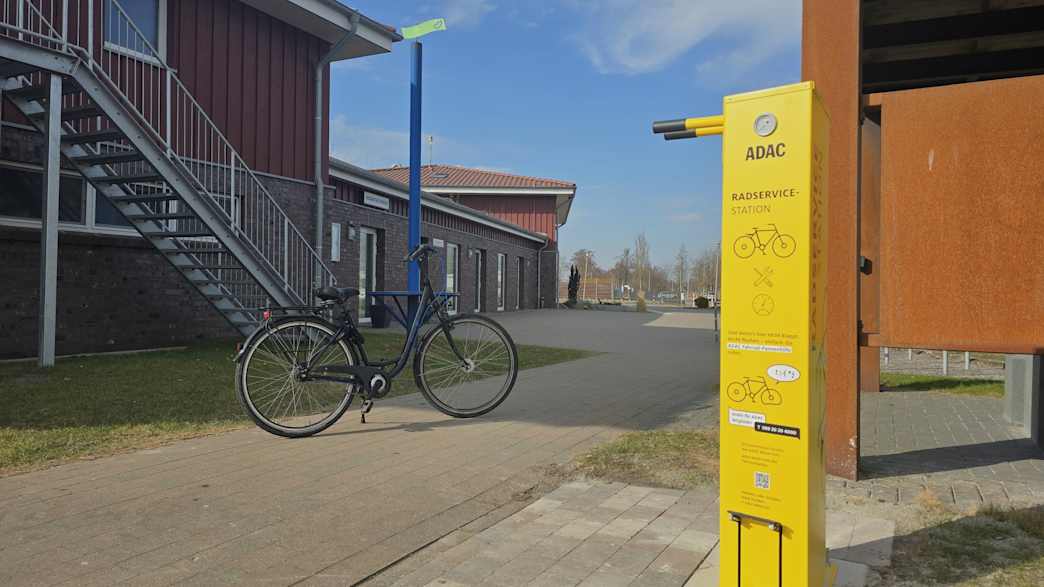 Ein Fahrrad steht auf einem asphaltierten Weg vor einem Haus. Auf der rechten Seite des Weges befindet sich eine gelbe Radservice-Säule.