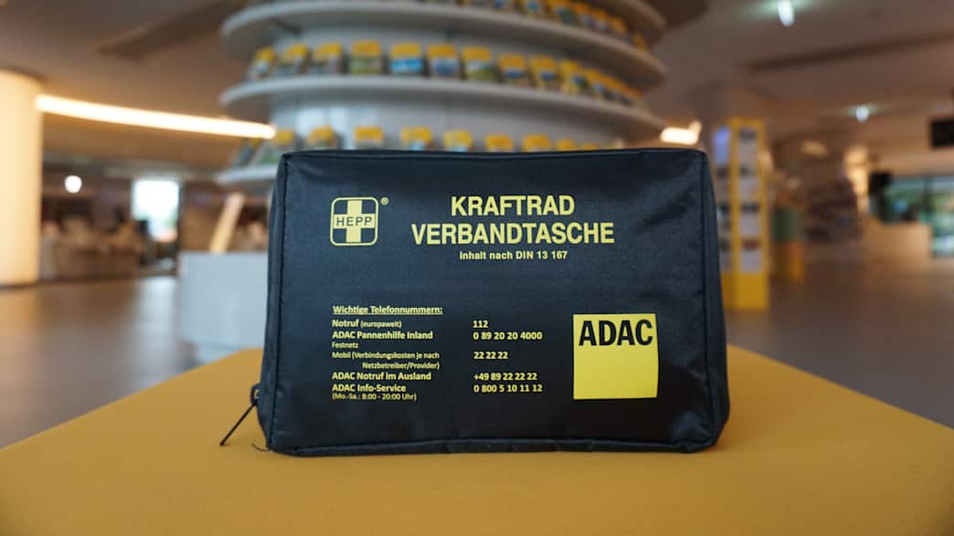 Eine Kraftrad-Verbandtasche steht auf einer gelben Oberfläche. Im Hintergrund ist eine ADAC Geschäftsstelle zu sehen.