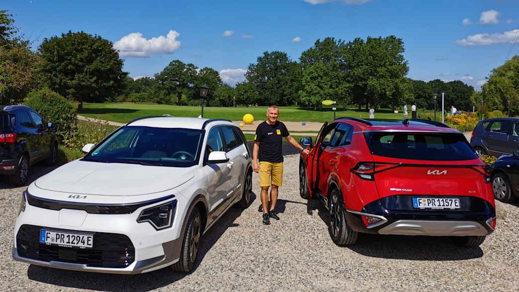 Ein Mann steht zwischen einem roten und einem weißen Kia Sportage auf einem Kiesparkplatz und zeigt wie man die Reichweite auf der Autobahn erweitert.
