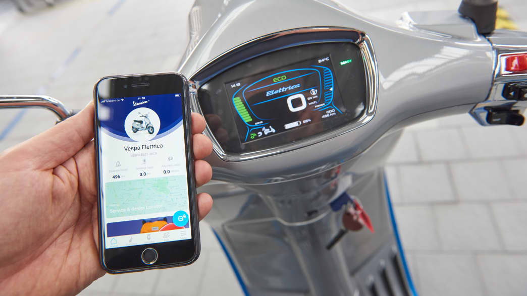 Ein Smartphone neben dem Display der Vespa Elletrica zeigt den Ladestand der Batterie an