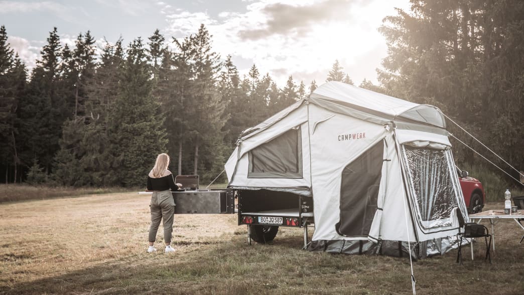 Eine Frau steht vor der Küchenzeile des Campwerk Economy Zeltanhängers auf einer Wiese im Wald