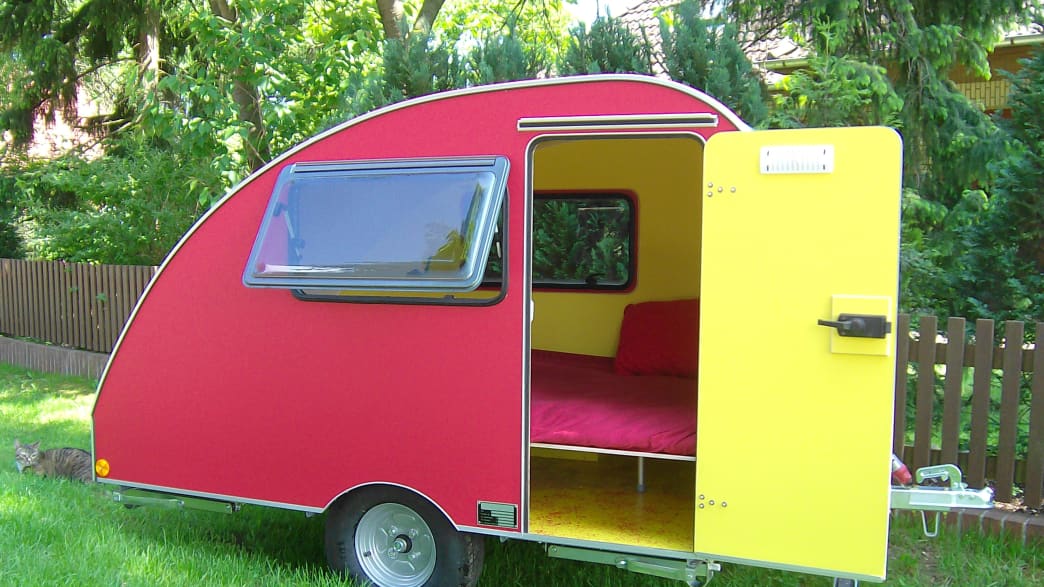 Das rot-gelbe, abgerundete Modell des Schneckenhaus Wohnmobilanhängers steht in einem Garten