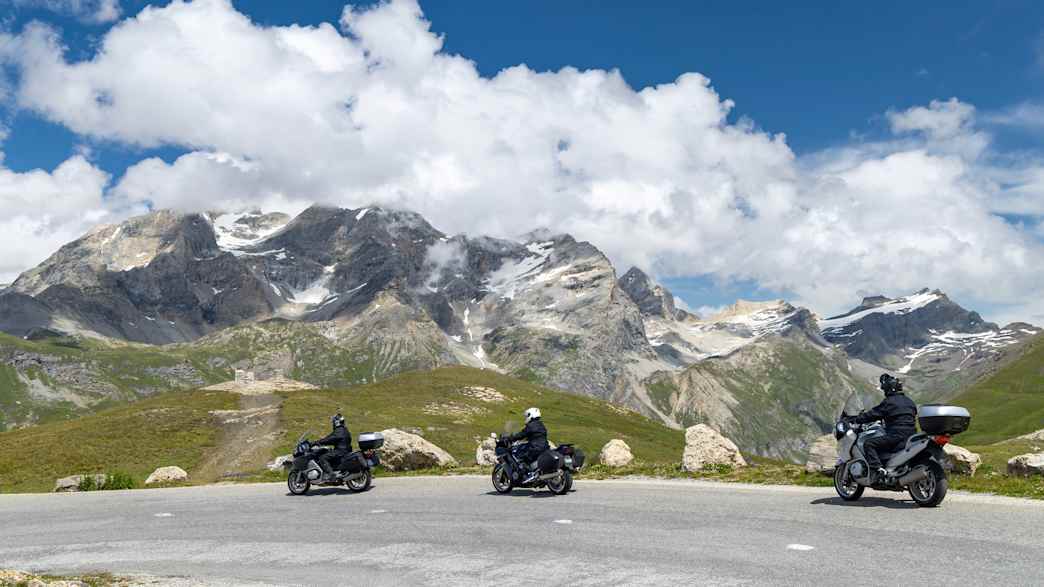 Motorradfahrer an der Passhöhe des Col de l’Iseran mit verschneiten Berggipfeln im Hintergrund.