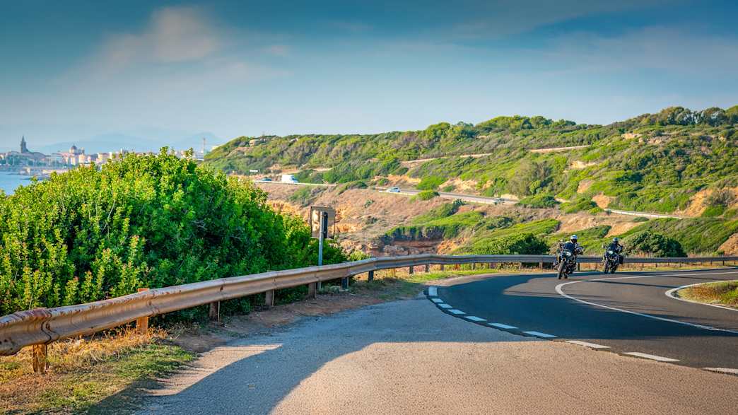 Motorradfahrer auf einer Straße auf Sardinien, im Hintergrund ist die Stadt Alghero zu sehen