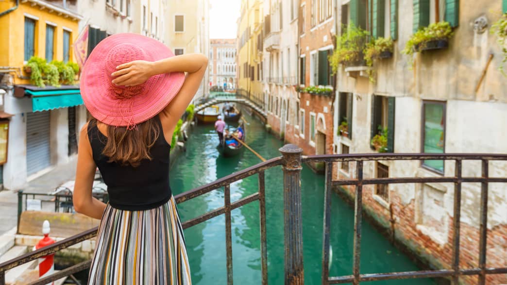 Elegante Frau mit rotem Sonnenhut genießt den Blick auf einen Kanal mit vorbeifahrender Gondel in Venedig, Italien
