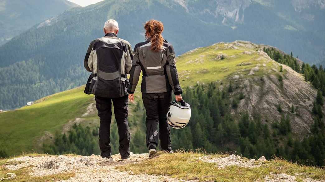 Ein Pärchen steht auf einem Hügel in Motorradkluft und bewundert die Berge