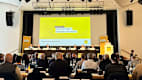 Bild des Podiums der Mitgliederversammlung. Im Vordergrund sieht man dies Besucher.