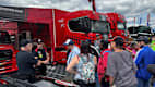 Eine Gruppe von Menschen steht vor roten Lastwagen auf einem Motorsport-Event. Einer der Menschen trägt ein schwarzes Hemd und spricht zu der Gruppe.
