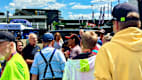 Eine Gruppe von Menschen steht bei einer Motorsport-Event-Location, während im Hintergrund Fahnen wehen. Der Himmel ist leicht bewölkt.