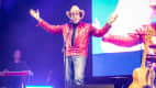 Country-Sänger, der live auf der Bühne auftritt, mit roter Lederjacke und Cowboyhut, vor einem lebhaften Publikum im Hintergrund.