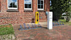 Vor einem Backsteingebäude mit drei Fenstern stehen Fahrradständer, eine gelbe Radservice-Station und eine E-Bike Ladesäule.