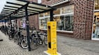Eine gelbe Säule steht vor einem überdachten Fahrradständer. Im Hintergrund ist ein Geschäft in der Fußgängerzone zu sehen.