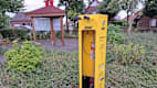 Eine gelbe Säule steht vor einem Beet mit Sträuchern. Im Hintergrund sind Häuser und eine Auskunftstafel zu sehen.