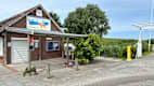 Links im Bild steht ein Kiosk mit einem überdachten Vorplatz. Daran anschließend stehen Bäume und Büsche und ein Feld. Vor dem Feld steht eine gelbe Radservice-Säule.