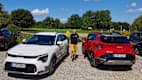 Ein Mann steht zwischen einem roten und einem weißen Kia Sportage auf einem Kiesparkplatz und zeigt wie man die Reichweite auf der Autobahn erweitert.