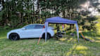 Ein Tesla Model Y steht auf einer Wiese, hinter ihm ist ein kleiner Pavillon aufgebaut mit Fahrrädern