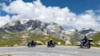 Motorradfahrer an der Passhöhe des Col de l’Iseran mit verschneiten Berggipfeln im Hintergrund.