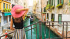 Elegante Frau mit rotem Sonnenhut genießt den Blick auf einen Kanal mit vorbeifahrender Gondel in Venedig, Italien