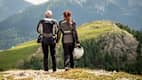 Ein Pärchen steht auf einem Hügel in Motorradkluft und bewundert die Berge