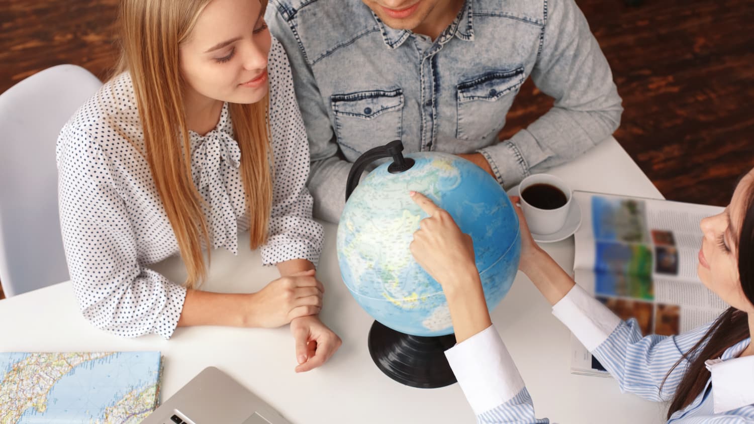 Young man and woman in a tour agency with a travel agent choosing destination on a globe looking curious smiling