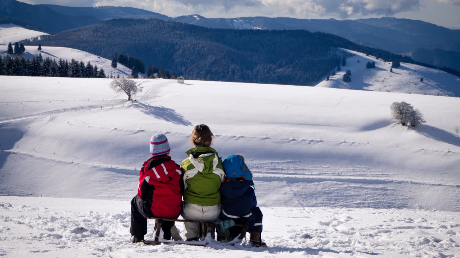 eine Frau und zwei Kinder sitzen auf einem Schlitten und schauen auf die verschneite Winterlandschaft vor ihnen