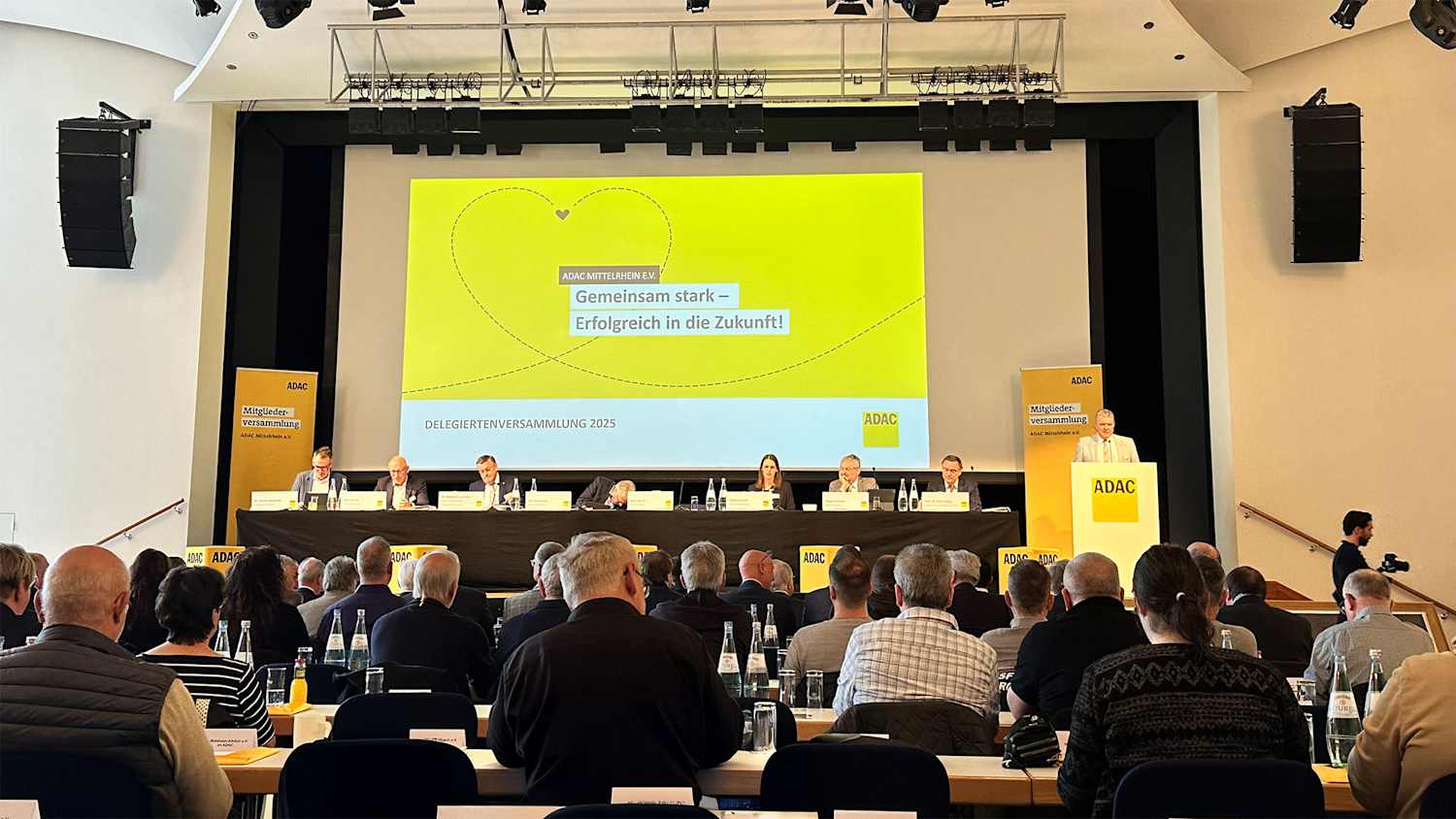 Bild des Podiums der Mitgliederversammlung. Im Vordergrund sieht man dies Besucher.