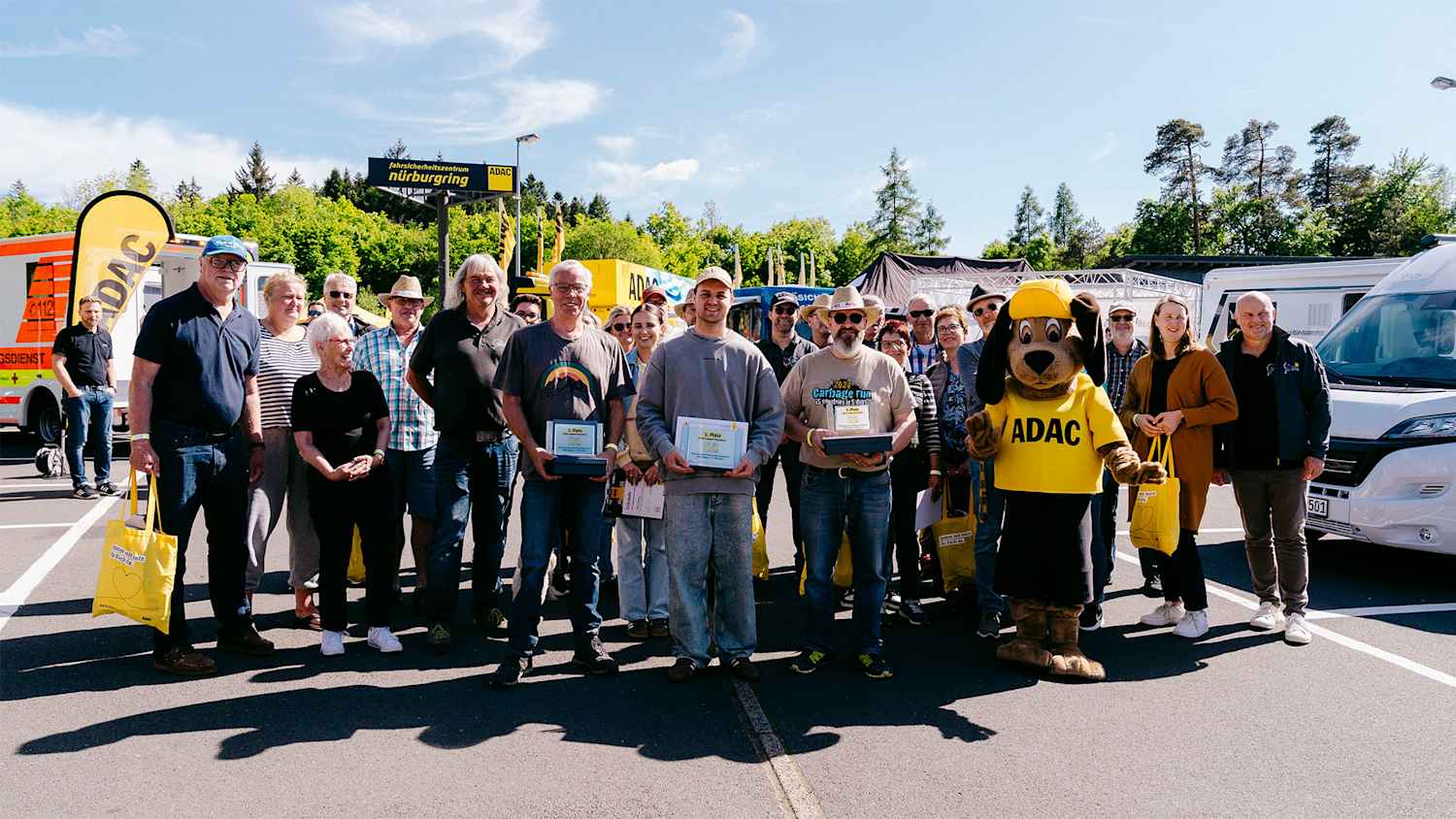 Eine Gruppe von Menschen posiert vor dem Fahrsicherheitszentrum Nürburgring. Neben ihnen steht ein ADAC-Maskottchen. Einige Personen halten Auszeichnungen in den Händen.