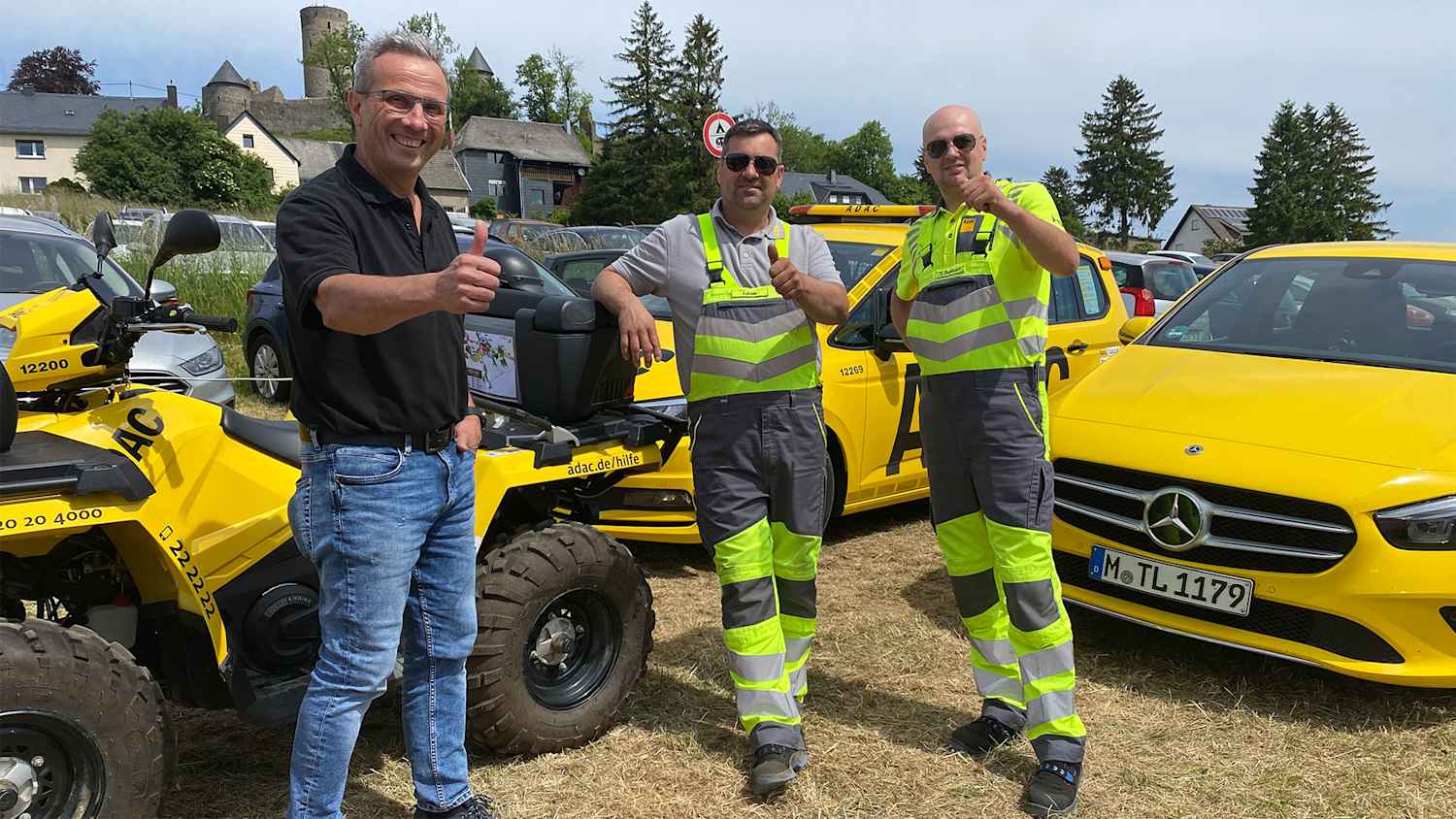 Drei Männer stehen vor gelben ADAC-Fahrzeugen und einem Quad, alle lächelnd und mit erhobenem Daumen. Im Hintergrund sind Häuser und Bäume zu sehen.