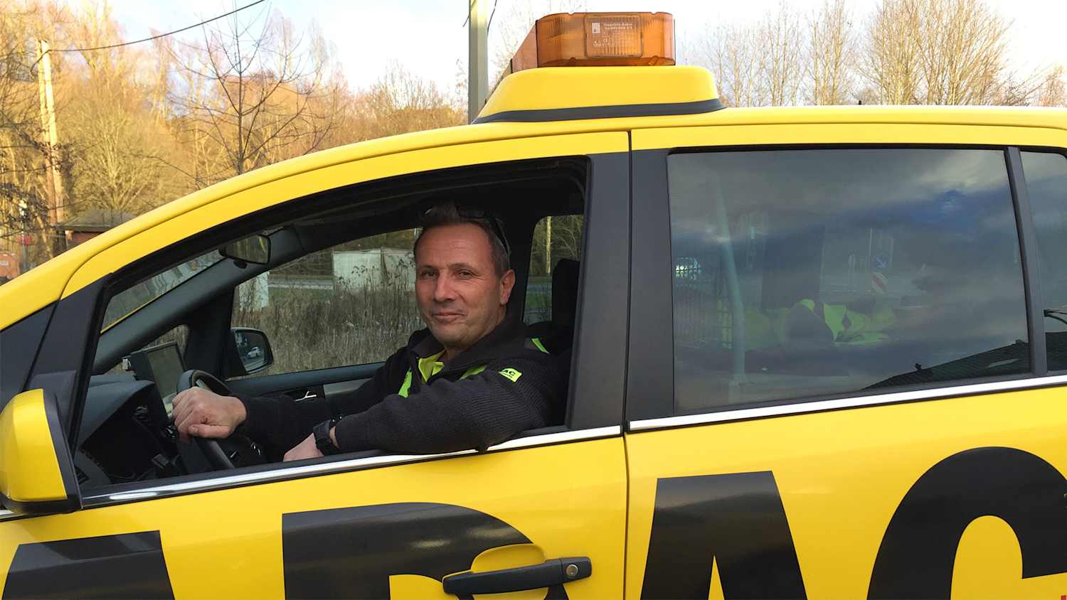 Eine Person sitzt am Steuer eines gelben ADAC-Fahrzeugs. Das Auto ist seitlich zu sehen und befindet sich im Freien bei Tageslicht.