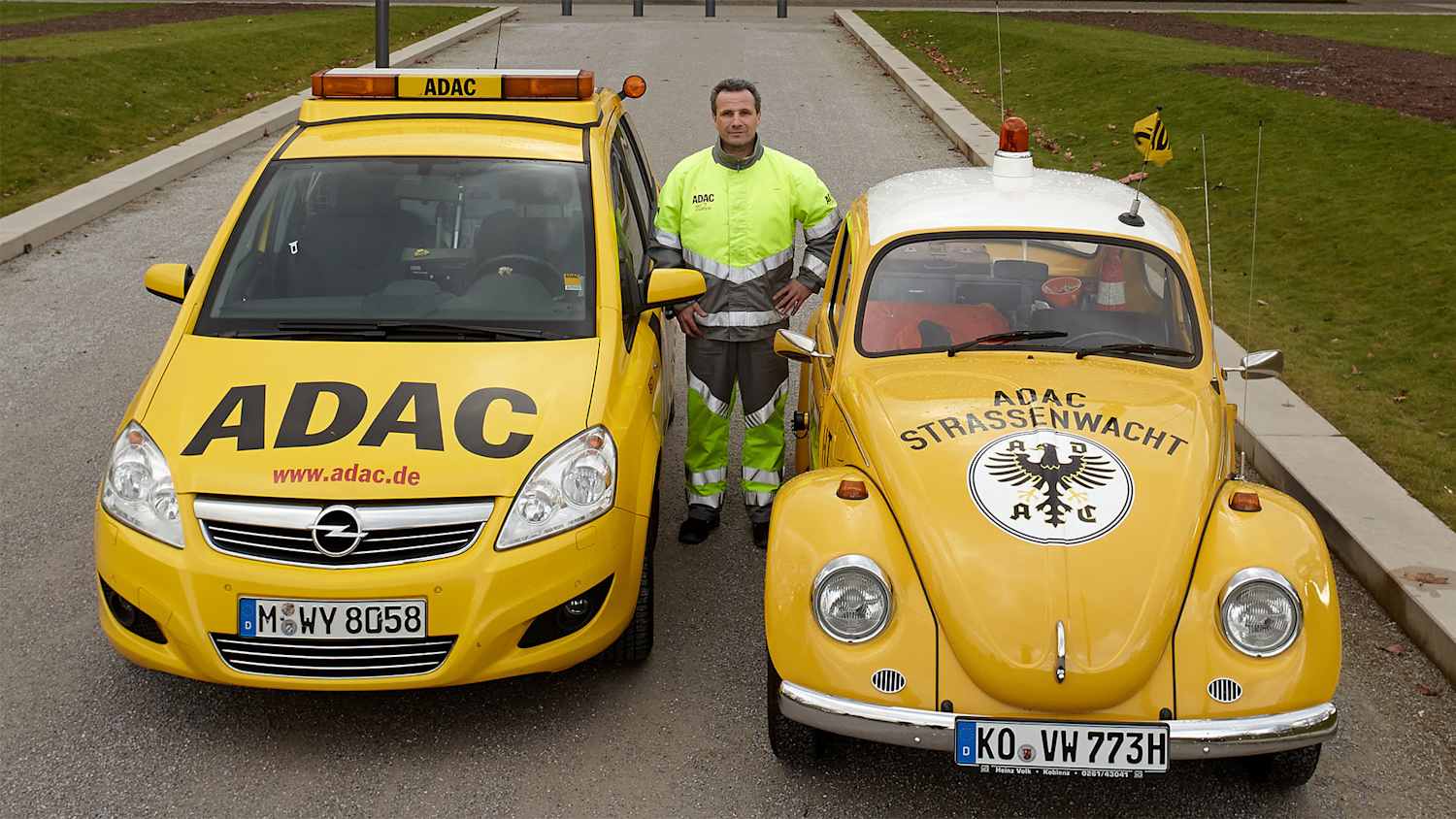 Zwei gelbe ADAC-Fahrzeuge, ein moderner Opel und ein klassischer VW Käfer, stehen nebeneinander vor einem Gebäude. Zwischen den Autos steht eine Person in gelber Warnkleidung.