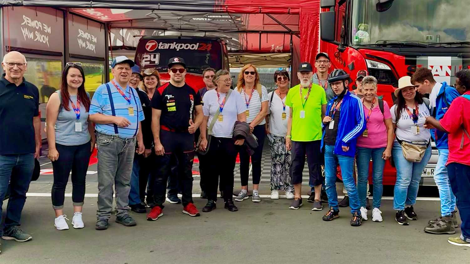 Eine Gruppe von Menschen steht vor einem roten Lastwagen, umgeben von Rennsport-Equipment. Der Himmel ist blau mit ein paar Wolken.